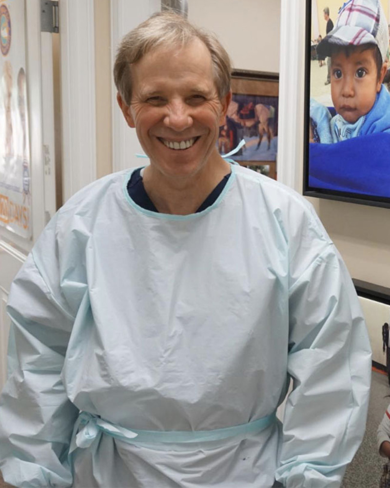 A smiling man in a light blue medical gown stands in an office, with a picture of a child in a cap in the background.